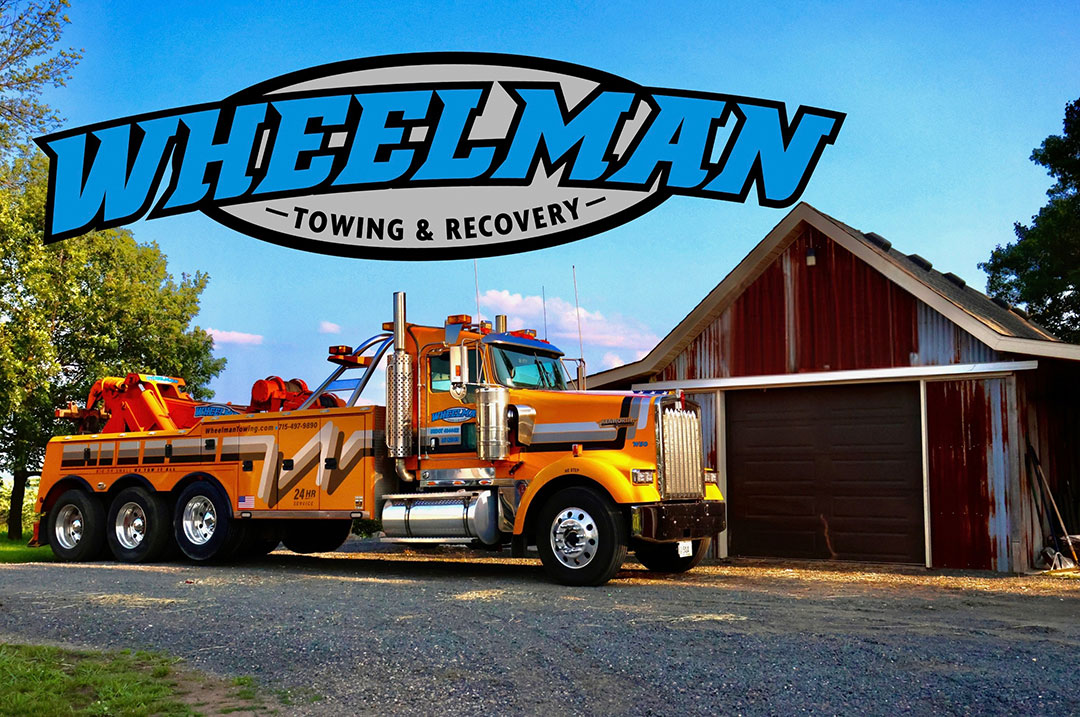 Orange Wheelman tow truck parked beside a red barn on a sunny day with the Wheelman logo above.